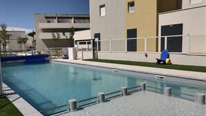 A swimming pool with a clear blue water, surrounded by a low concrete deck. There are metal railings along the pool's edge. In the background, there is a modern building with multiple windows and doors. - La Dune by Interhome (Valras Plage)