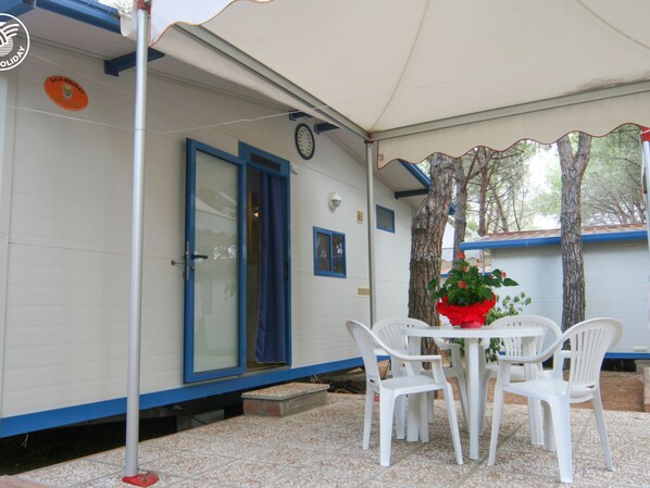 Un espace extérieur abrité avec une table et des chaises blanches, une plante en pot et un bâtiment doté d’une porte et de fenêtres bleues.