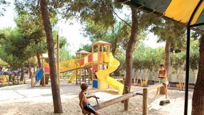 Une aire de jeux avec un toboggan et des structures d’escalade, entourée d’arbres et d’une zone sablonneuse.