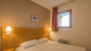 A hotel room with a wooden headboard, two bedside lamps, a window with red curtains, and a framed picture on the wall.