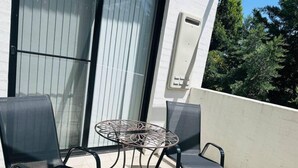 Un espace extérieur aménagé avec une table ronde et quatre chaises, une porte-fenêtre coulissante et un mur blanc sur lequel est fixé un petit appareil de climatisation.