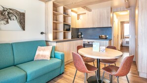 Una cocina americana moderna con microondas, cafetera y tostadora. Hay un sofá color turquesa con un cojín decorativo, una mesa de comedor redonda con cuatro sillas y un techo de madera con iluminación empotrada.
