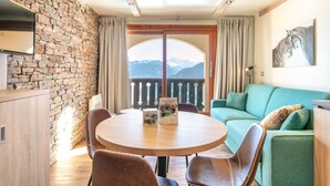 Una habitación acogedora con pared de piedra, techo de madera, una mesa redonda con sillas, un sofá verde y un balcón con vistas a la montaña.