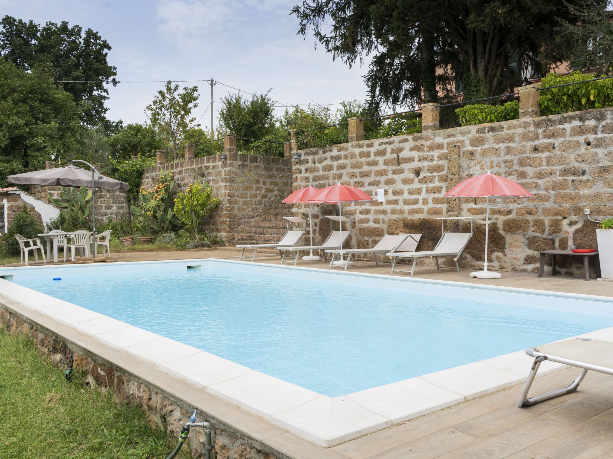 A swimming pool with lounge chairs and umbrellas, surrounded by a stone wall and greenery.