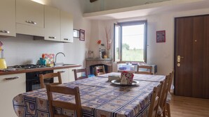 Un coin repas avec une table dressée pour quatre personnes, des chaises en bois, une cuisine avec des meubles blancs, une cuisinière et une fenêtre donnant sur un paysage verdoyant.