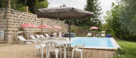 A poolside area with lounge chairs and umbrellas, a stone wall, and a grassy area with trees.