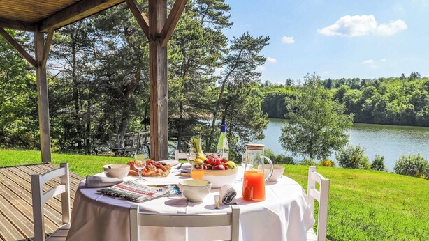 Mesa al aire libre con vista a un lago y un bosque, mostrando una comida con frutas, una botella y un vaso de jugo.