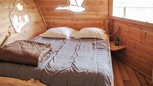 Un dormitorio de cabaña de madera con una cama, almohadas, una lámpara colgante y una pequeña mesa con un farol.