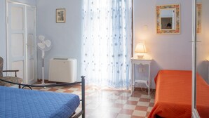 Ein Schlafzimmer mit zwei Betten, einem Ventilator, einer Lampe, einem Spiegel und einem Fenster mit floralen Vorhängen.