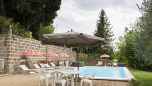 Ein Poolbereich mit Liegestühlen und Sonnenschirmen, eine Steinmauer und eine begrünte Fläche mit Bäumen.