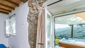 A room with a stone wall, a window with curtains, a wicker chair, and a view of a hot tub and mountains.