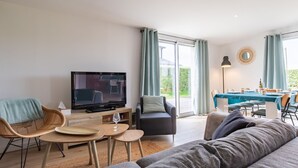Un salon moderne doté d’un téléviseur à écran plat, de sièges confortables, d’un coin repas avec une table et des chaises, et de grandes fenêtres offrant une vue sur l’extérieur.