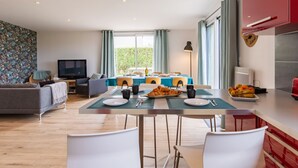 Un salon moderne avec un canapé et un téléviseur, un coin repas avec une table dressée pour deux personnes, et une cuisine avec des placards rouges et une grande fenêtre.