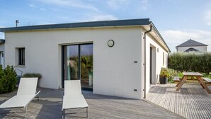 Une maison moderne avec un toit plat, une porte-fenêtre coulissante et une terrasse en bois équipée de deux chaises longues.