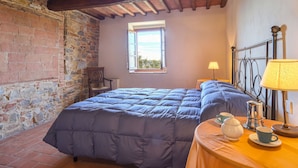 Ein Schlafzimmer mit Bett, Fenster, Stuhl, Tisch mit Lampe, Kaffeemaschine und Tasse.