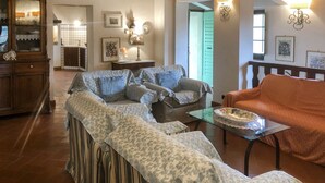 Un salón acogedor con un sofá, sillones y una mesa de centro de vidrio. Se observa un mueble de madera y una mesa de comedor con un plato.