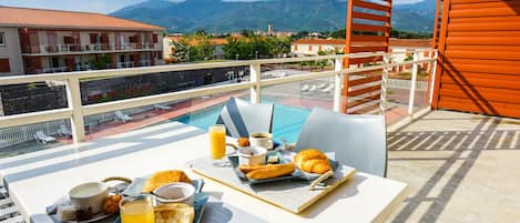 A balcony with a table set for breakfast, overlooking a swimming pool and mountains.