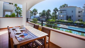 En balkon med et træbord, stole og udsigt til en swimmingpool og bygninger.