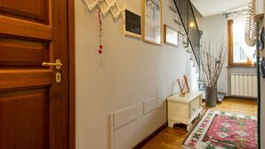 A hallway with a wooden door, a white wall with framed pictures, a small white side table with a vase, a staircase, and a radiator. - La Rondine (LUU280) by Interhome (Lucca)