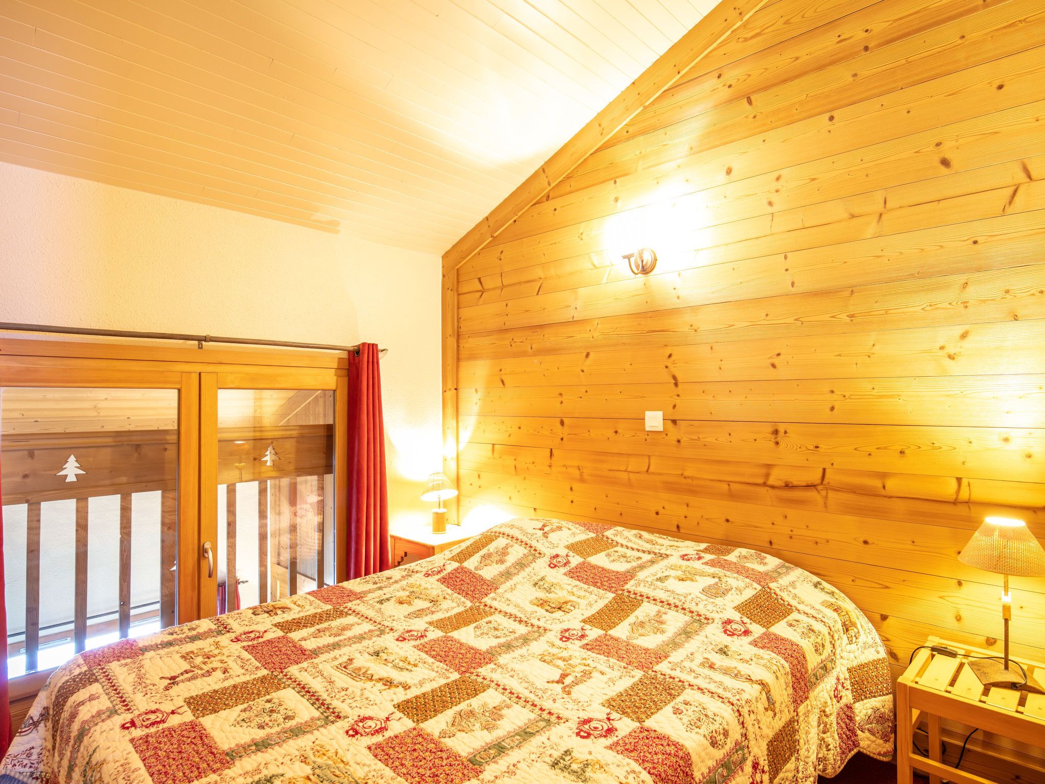 A cozy room with a wooden ceiling, a large bed with a patterned quilt, a sliding glass door, and two bedside lamps.