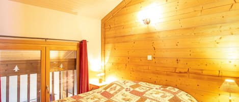 A cozy room with a wooden ceiling, a large bed with a patterned quilt, a sliding glass door, and two bedside lamps.