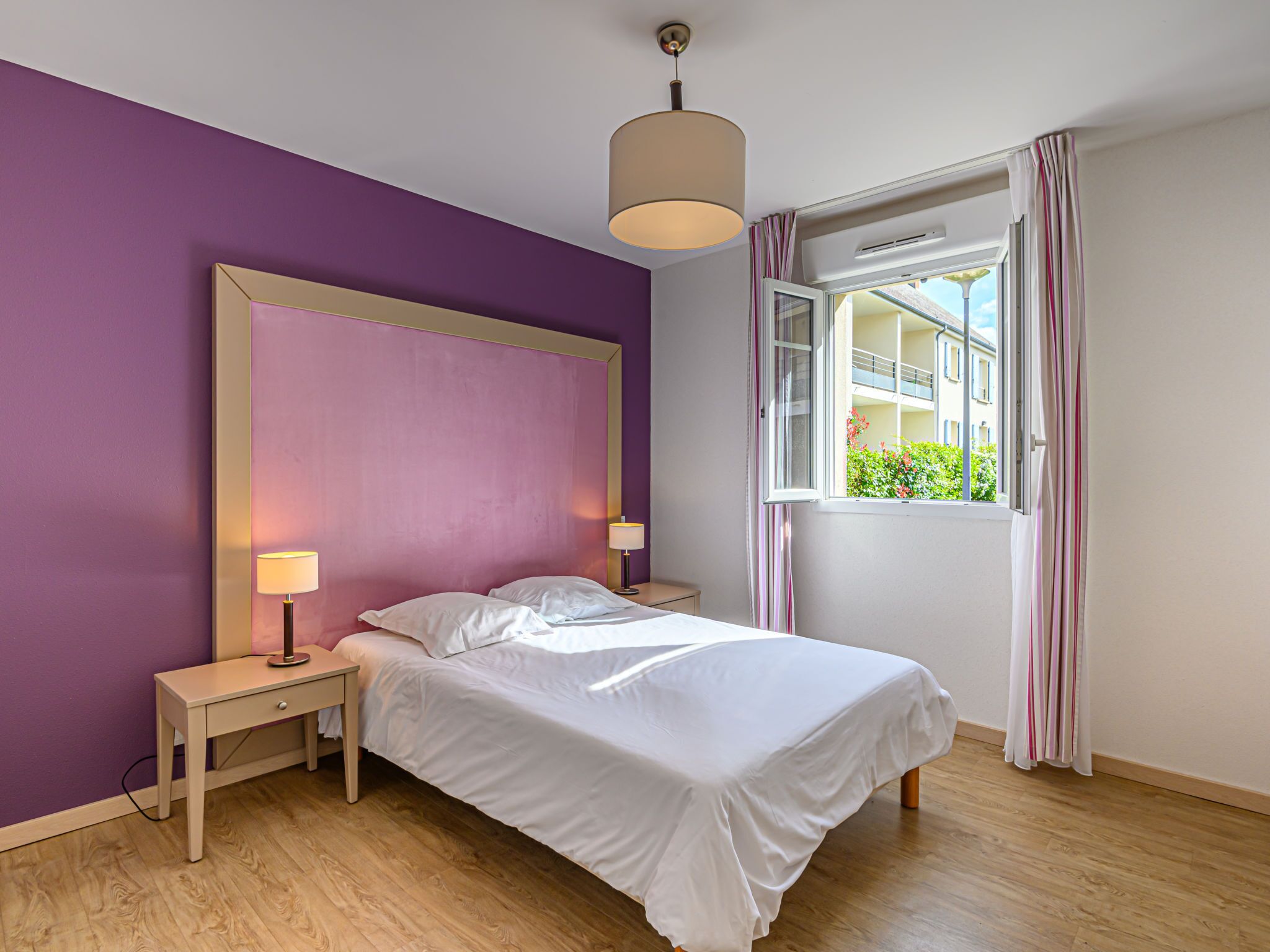 Une chambre à coucher avec un lit, des tables de chevet, une lampe, une fenêtre avec des rideaux et une vue sur les bâtiments à l’extérieur.