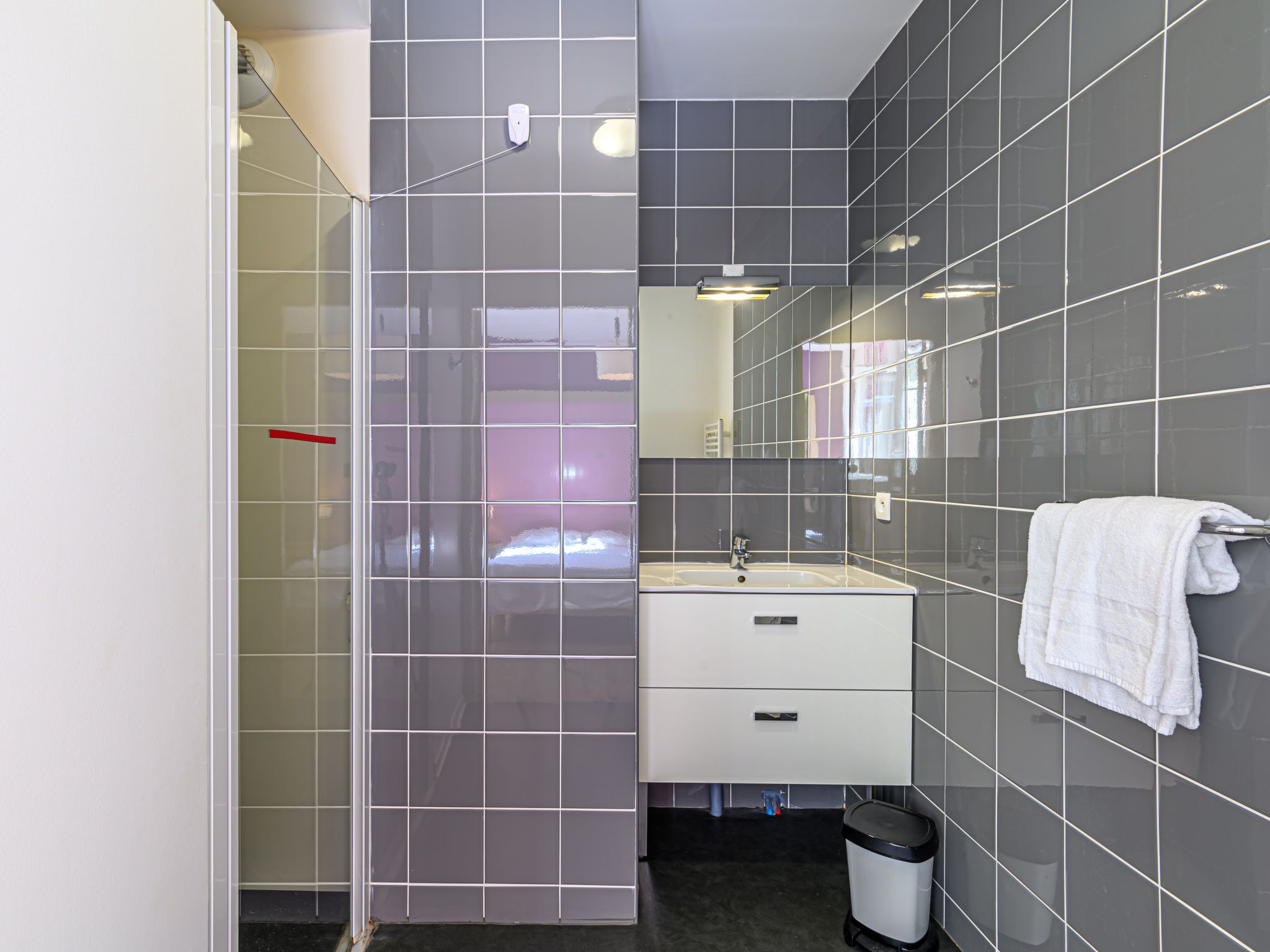 Une salle de bain moderne avec une douche à l’italienne en verre, un lavabo blanc avec un miroir au-dessus et une serviette accrochée au mur.