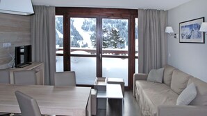 A modern hotel room with a wooden dining table, a sofa, and a TV on a wooden cabinet. Large windows offer a view of a snowy mountain landscape.