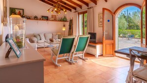 Uma sala de estar aconchegante com teto de madeira, mesa de centro de vidro, poltronas com almofadas verdes, televisão e vista para o exterior através de uma porta de vidro.