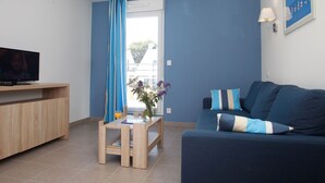Un salon moderne avec un téléviseur posé sur un meuble en bois, une petite table en bois sur laquelle se trouve un verre de jus d’orange, un canapé bleu agrémenté de coussins à rayures et une fenêtre ornée de rideaux bleus.