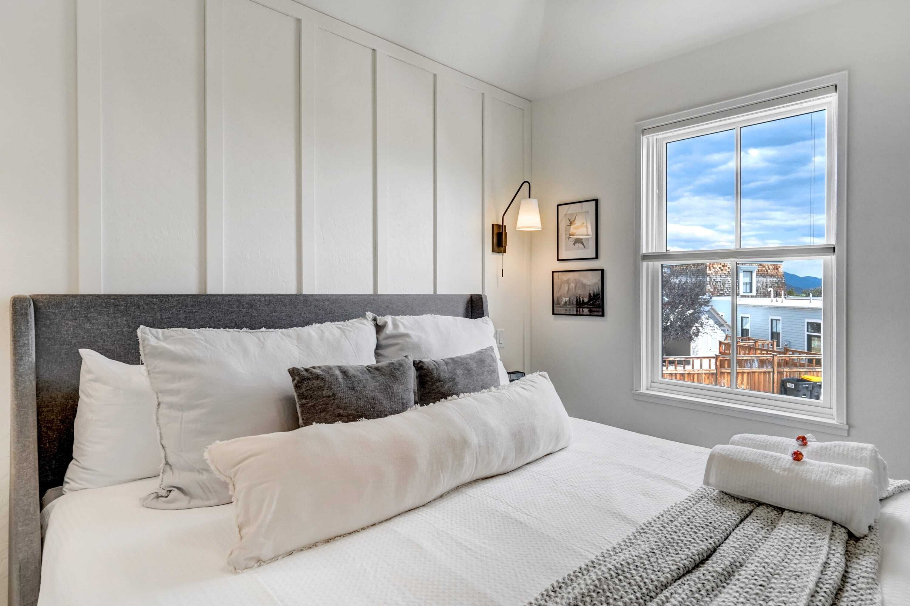 A neatly made bed with white and gray bedding, a gray headboard, and a view of a blue building through a window.