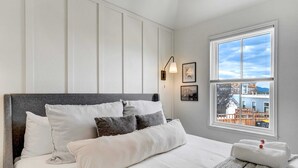 Una cama bien tendida con ropa de cama blanca y gris, un cabecero gris y vistas a un edificio azul a través de una ventana.
