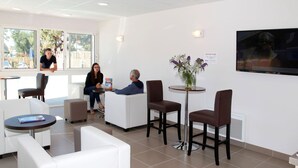 Un espace d’attente moderne avec des canapés blancs, une petite table ornée d’un vase de fleurs et un téléviseur fixé au mur.