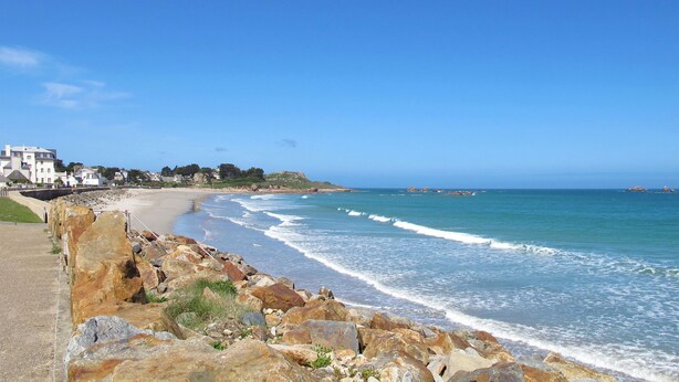A coastal view with a rocky shoreline, a sandy beach, and buildings in the background. - Odalys Domaine des Roches Jaunes by Interhome (Plougasnou)