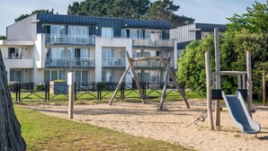 A playground with a slide and swings, surrounded by a fence, with a building in the background. - Odalys Domaine des Roches Jaunes by Interhome (Plougasnou)