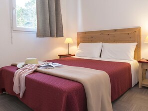 Un lit bien fait, recouvert d’une couverture rouge et beige, avec deux oreillers blancs et une tête de lit en bois. Une table de chevet avec une lampe et un livre est placée à côté du lit. Un chapeau et des lunettes sont posés sur le lit.