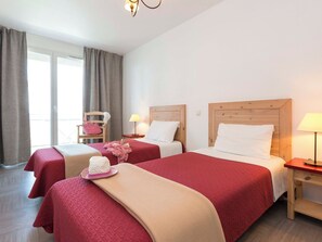 Habitación de hotel con dos camas, cada una con cabecero de madera, una mesita de noche con lámpara y una silla con un bolso rosa.