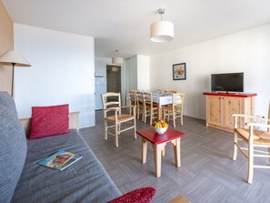 Un salon moderne avec un canapé gris, des coussins rouges, une table à manger avec des chaises, un téléviseur posé sur un meuble en bois, et un couloir menant à la cuisine.