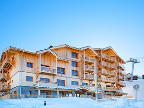 Un immeuble en bois à plusieurs étages, doté de balcons et d’une base en pierre, avec un télésiège visible en arrière-plan.