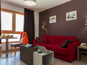 Ein modernes Wohnzimmer mit einem roten Sofa, einem Laptop auf einem Beistelltisch, einem Esstisch mit Frühstücksgebäck und zwei gerahmten Bildern an der Wand.