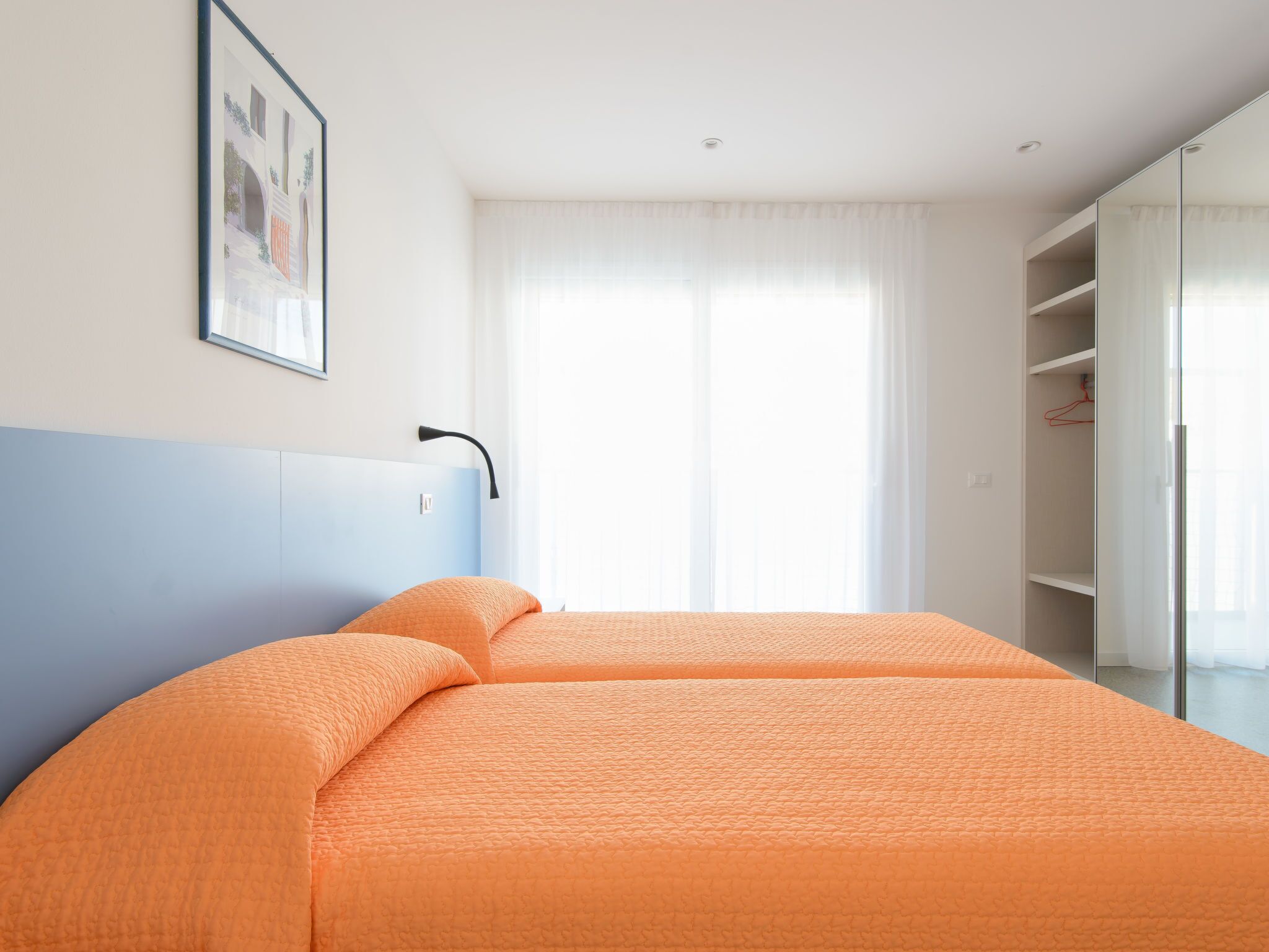 A bedroom with a bed, a nightstand, a wall-mounted lamp, a window with curtains, and a framed picture on the wall.