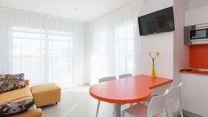 A modern living room with a sofa, a dining table, and chairs. A flat-screen TV is mounted on the wall above the table.