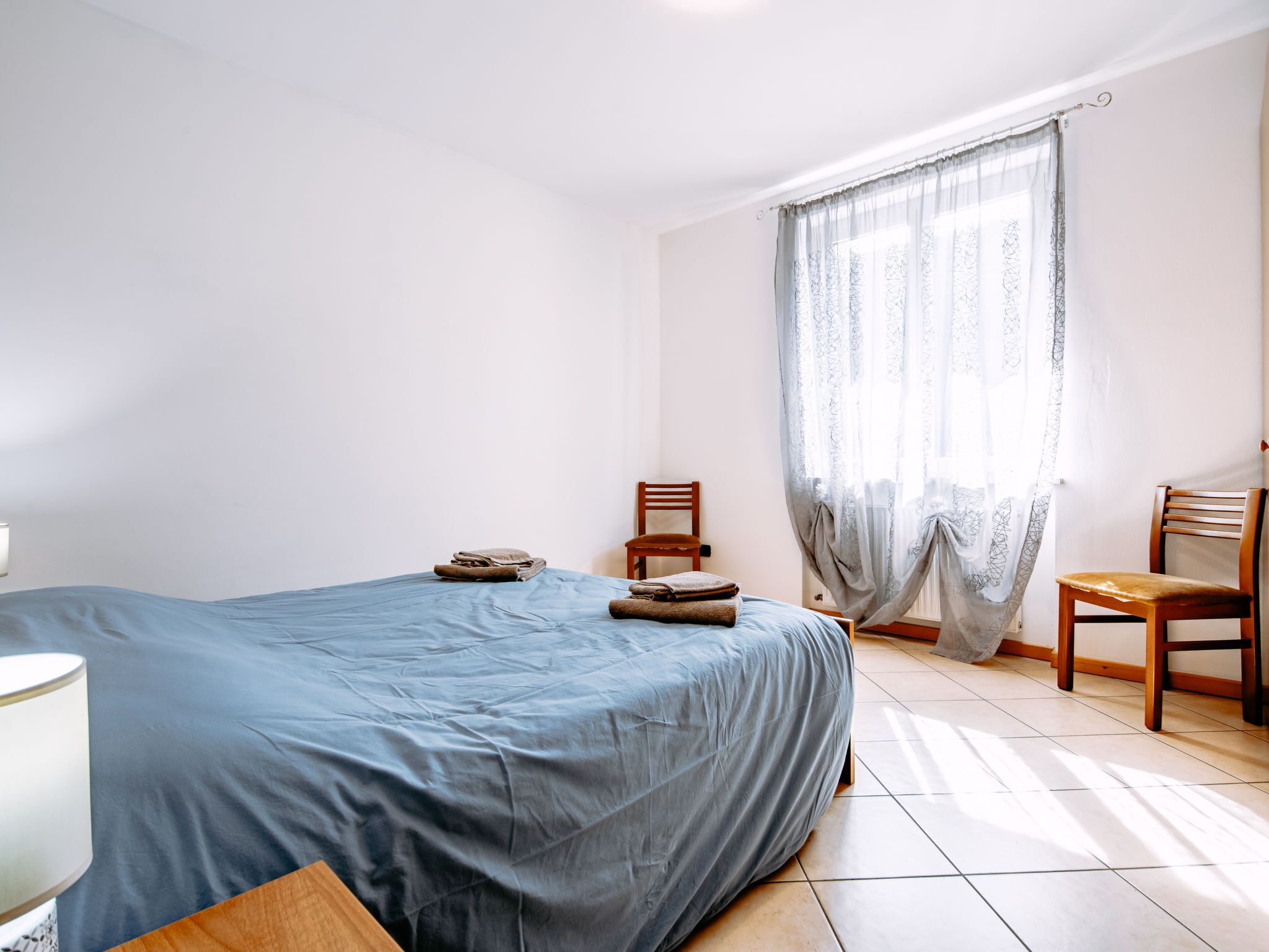 Une chambre à coucher avec un lit, deux chaises, une lampe et une fenêtre avec des rideaux.