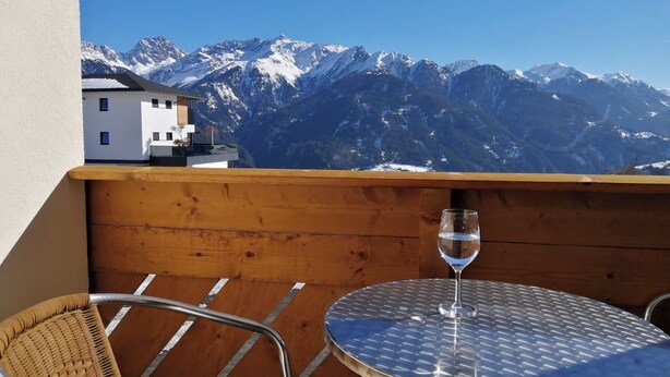 Un balcó amb vista a muntanyes nevades, una cadira de vimetla i una taula amb un got d'aigua.