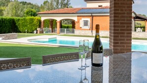 A poolside table with a bottle of wine and glasses, a brick house with a tiled roof, and a clear blue sky. - Appendino by Interhome (Asti)