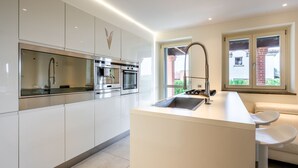 A modern kitchen with white cabinetry, stainless steel appliances, a central island with a sink, and a window with a view of a brick building. - Appendino by Interhome (Asti)