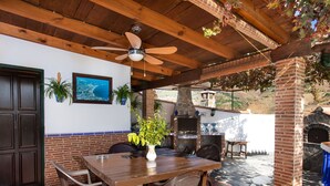 An outdoor patio with a wooden table and chairs, a ceiling fan, and a brick oven.