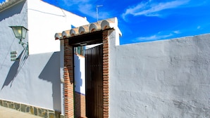 A narrow alleyway with a white building on one side and a stone wall on the other. A wooden door with a metal gate is visible in the center.