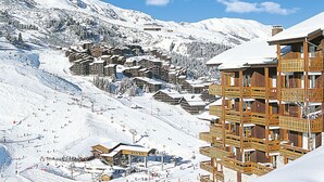 Un centro turístico en la montaña cubierto de nieve, con edificios de madera, pistas de esquí y un telesilla.
