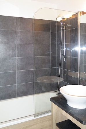 Un baño moderno con ducha envuelta en vidrio, un lavamanos blanco y paredes revestidas de baldosas oscuras.